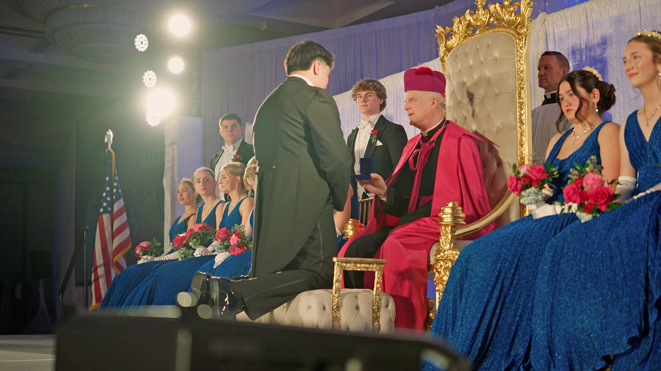 Young man in tuxedo kneels before bishop